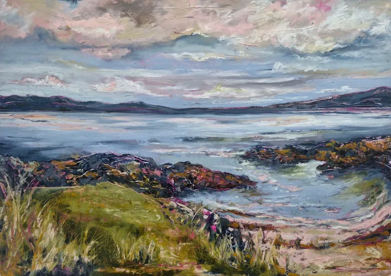 Rocks and green grass in the foreground, looking across a bay of pale water to distant hills under a pink tinged sky